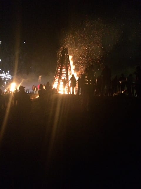 A bonfire lights a dark night.  A crowd is silhouetted against the light.  Fireworks are going off in the background.