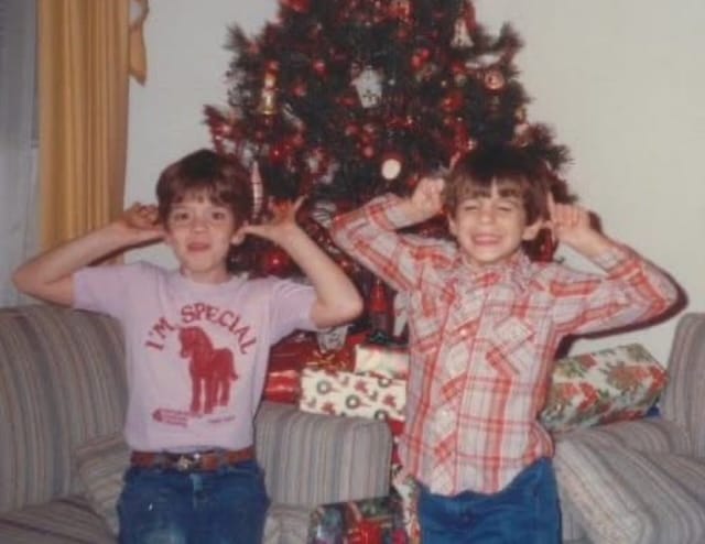 A grainy photo of two children posing in front of a decorated Christmas tree in a 1980s living room.