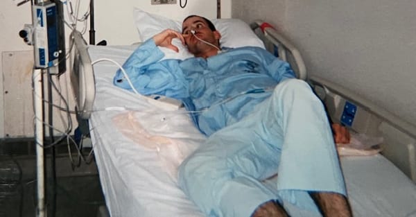 A young soldier in blue pajamas sitting in a hospital bed.  There's a feeding tube in his nose leading to an IV pump.