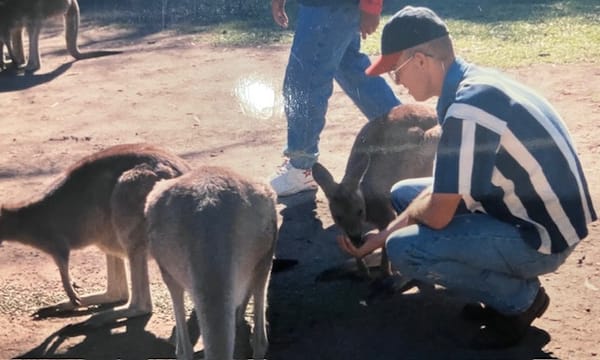 A white man in blue jeans, a blue and white striped shirt and a blue and red baseball cap is hand feeding a wallaby