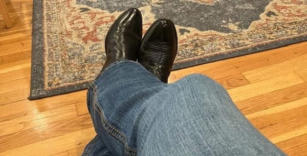 A pair of  blue jeans and black, pointed toe, western, cowboy boots on a light brown hardwood floor.